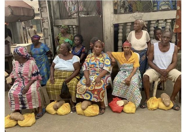 Older women and a man sit together outdoors with yellow food bags and tubers in front of them.