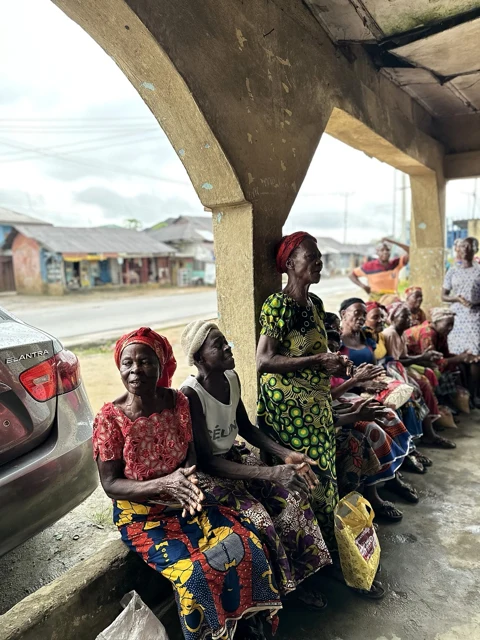 Older women sit and stand together beside a pillar, with bags of supplies at their feet.