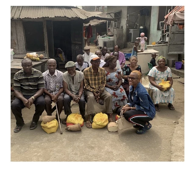 Older men and women gather outdoors with yellow bags and tubers, beside a woman crouching in front.