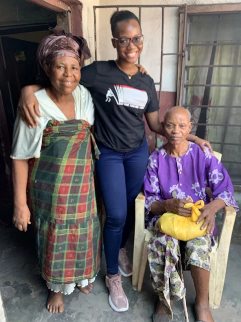 A woman stands beside two older women, one seated with a yellow bag.