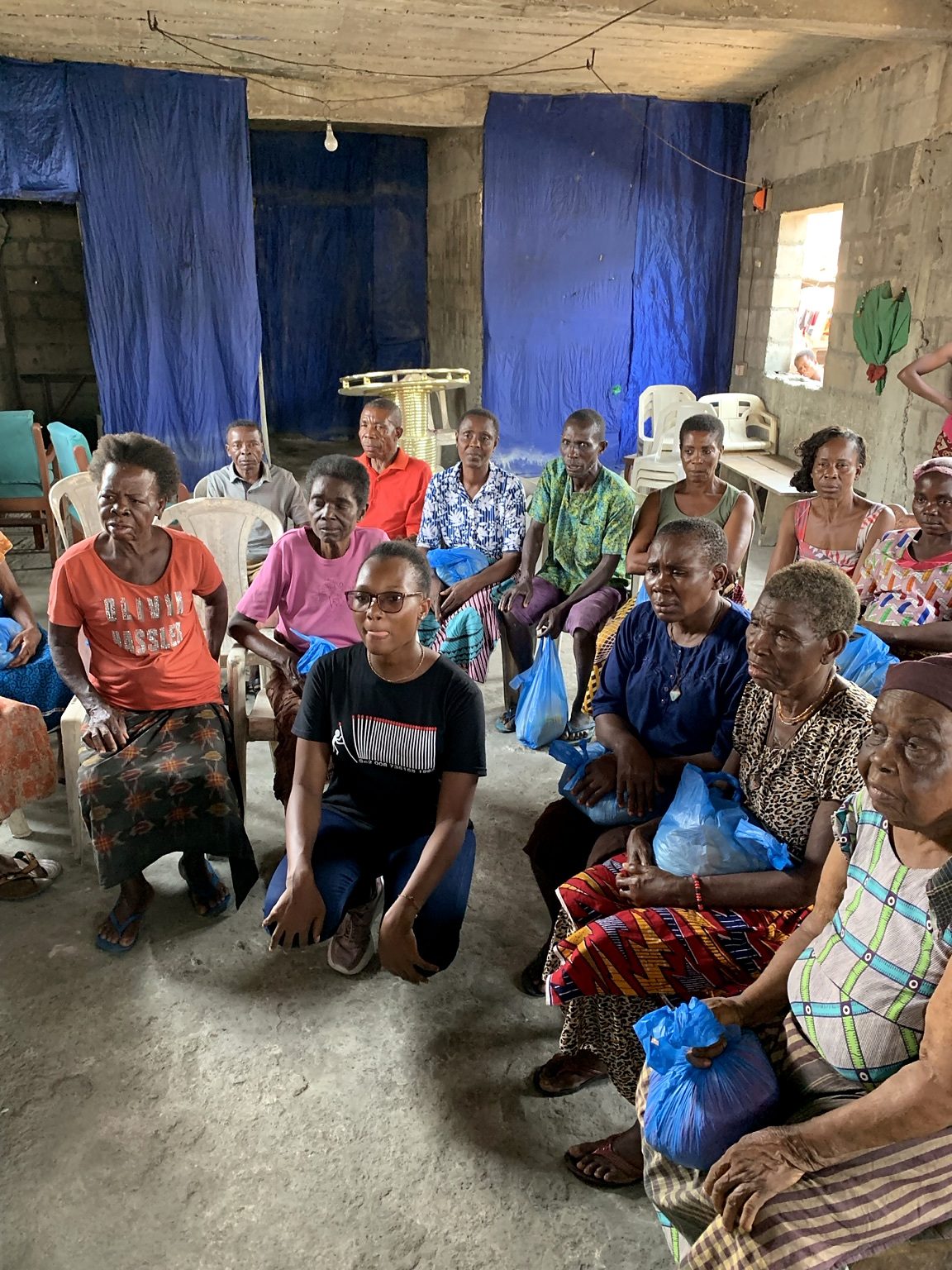 A group of older women and men sits together with blue bags, with a woman kneeling in front.