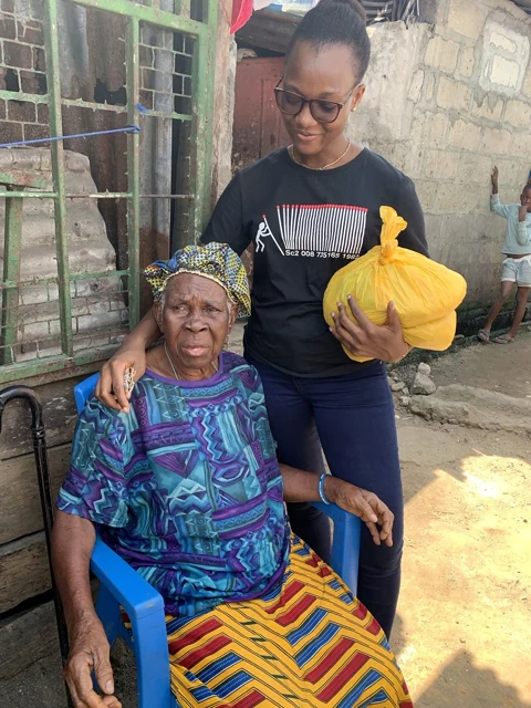 A woman holding a yellow bag rests her arm around an older woman seated in a blue chair.