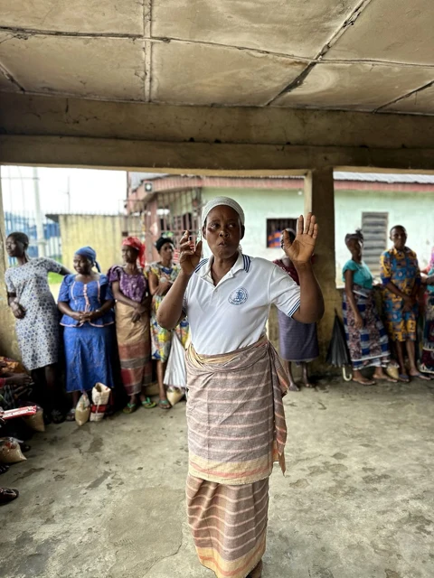 A woman in a white polo shirt gestures while older women stand and sit behind her.